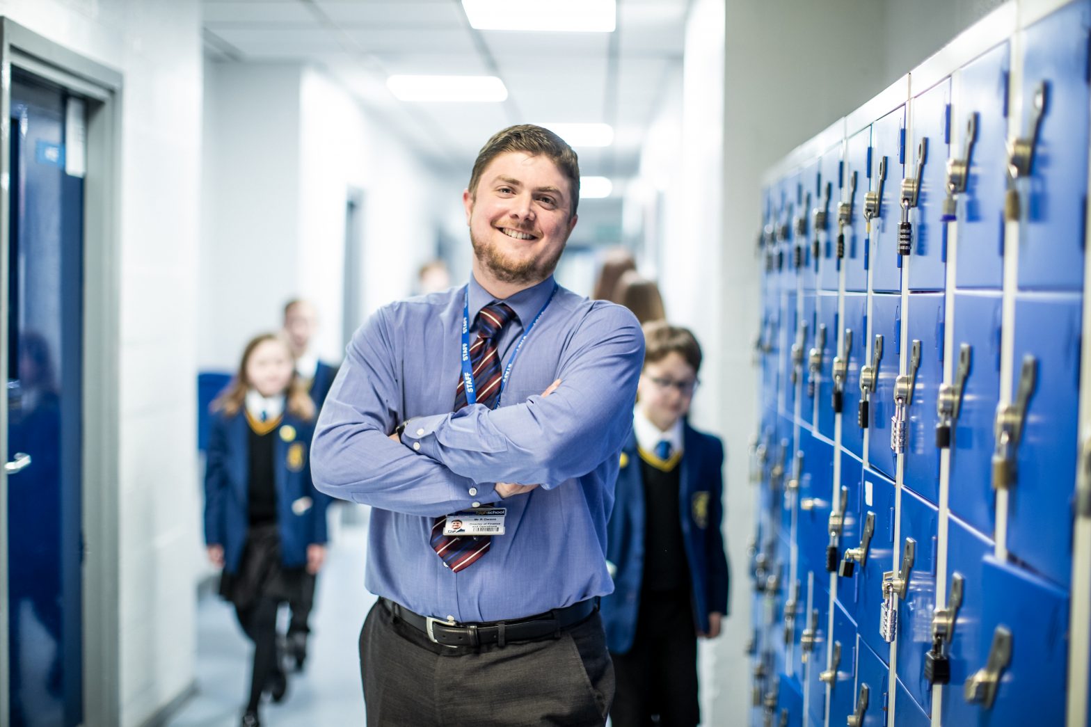 School teacher shown in the foreground smiling with his arms crossed and children walking behind him slightly blurred