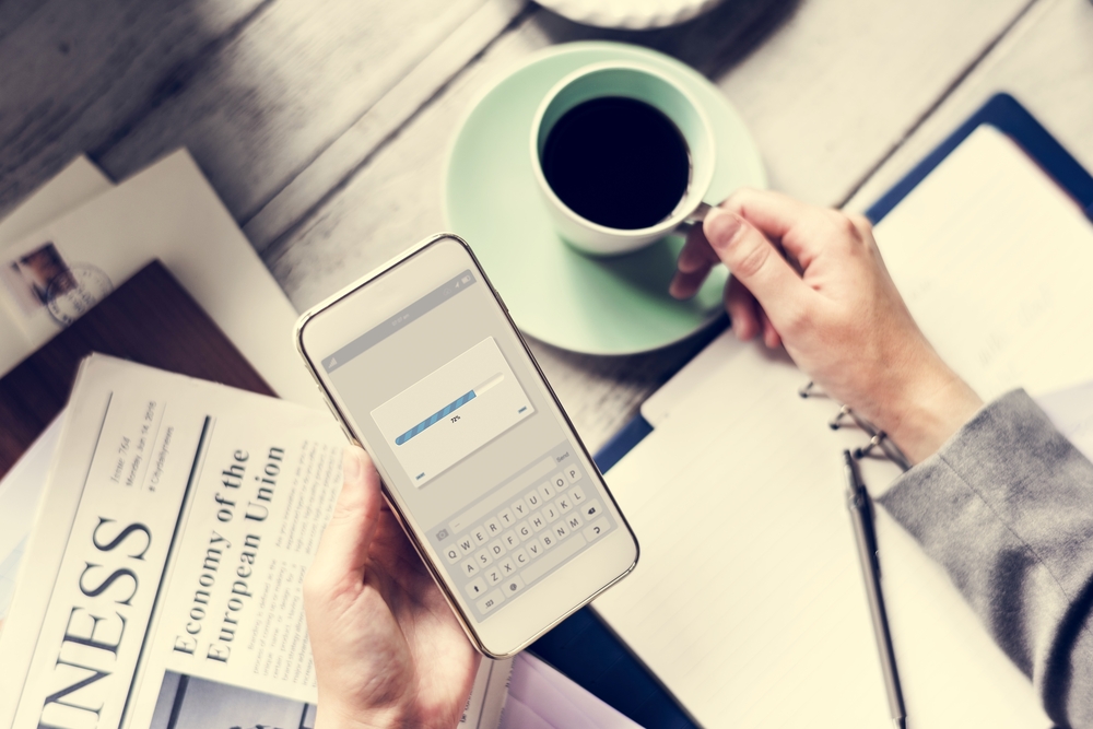 Hands Holding Downloading Mobile Phone with Coffee Cup and Newspaper