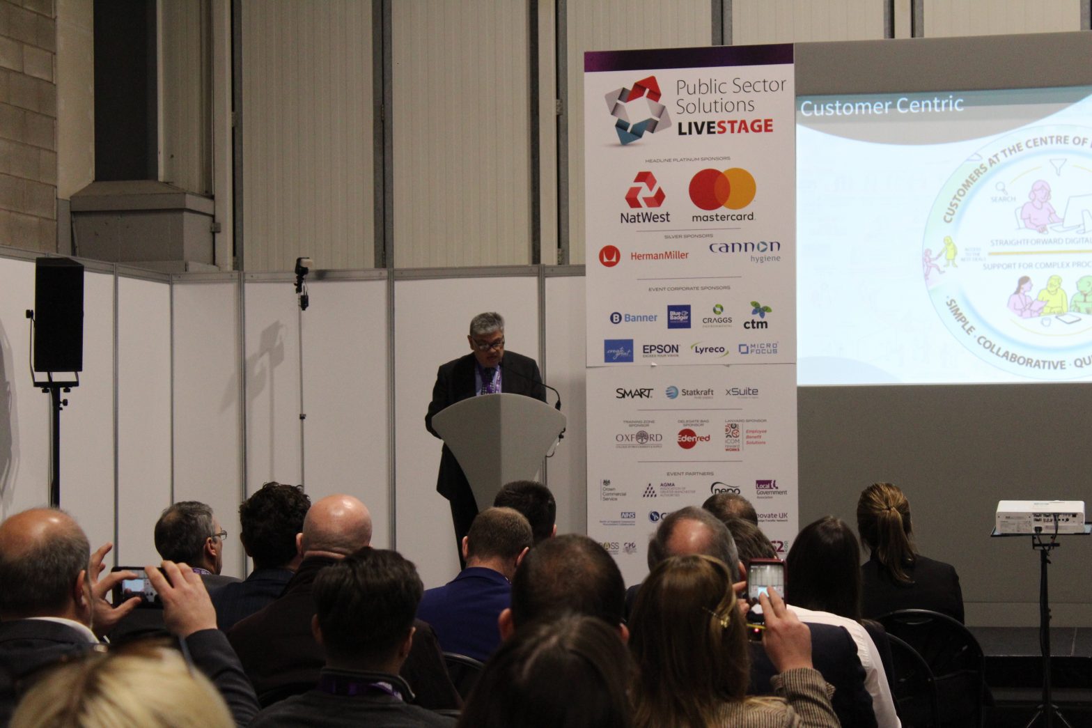 Simon Tse presents to a busy room at Procurex National. Behind him is a presentation screen and branding from different partner organisations.