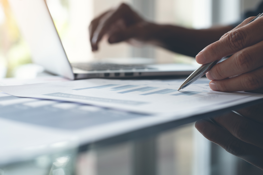 Hand holding a pen pointing to a graph printed on a piece of paper with a hand hovering over a laptop in the background which is slightly blurred