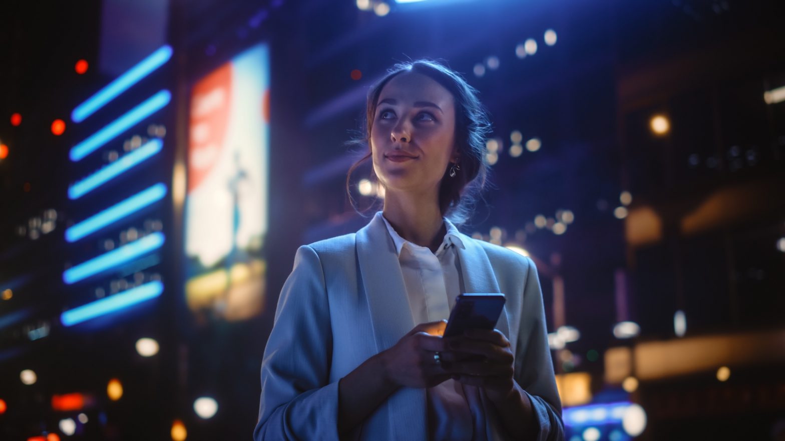 Nightime image of a woman in a city with buildings behind her. She is holding a mobile phone and looking to the upper left hand side of the image.