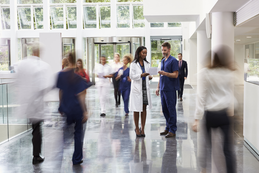 Staff In busy lobby area of modern hospital with 2 doctors shown talking in focus in the middle