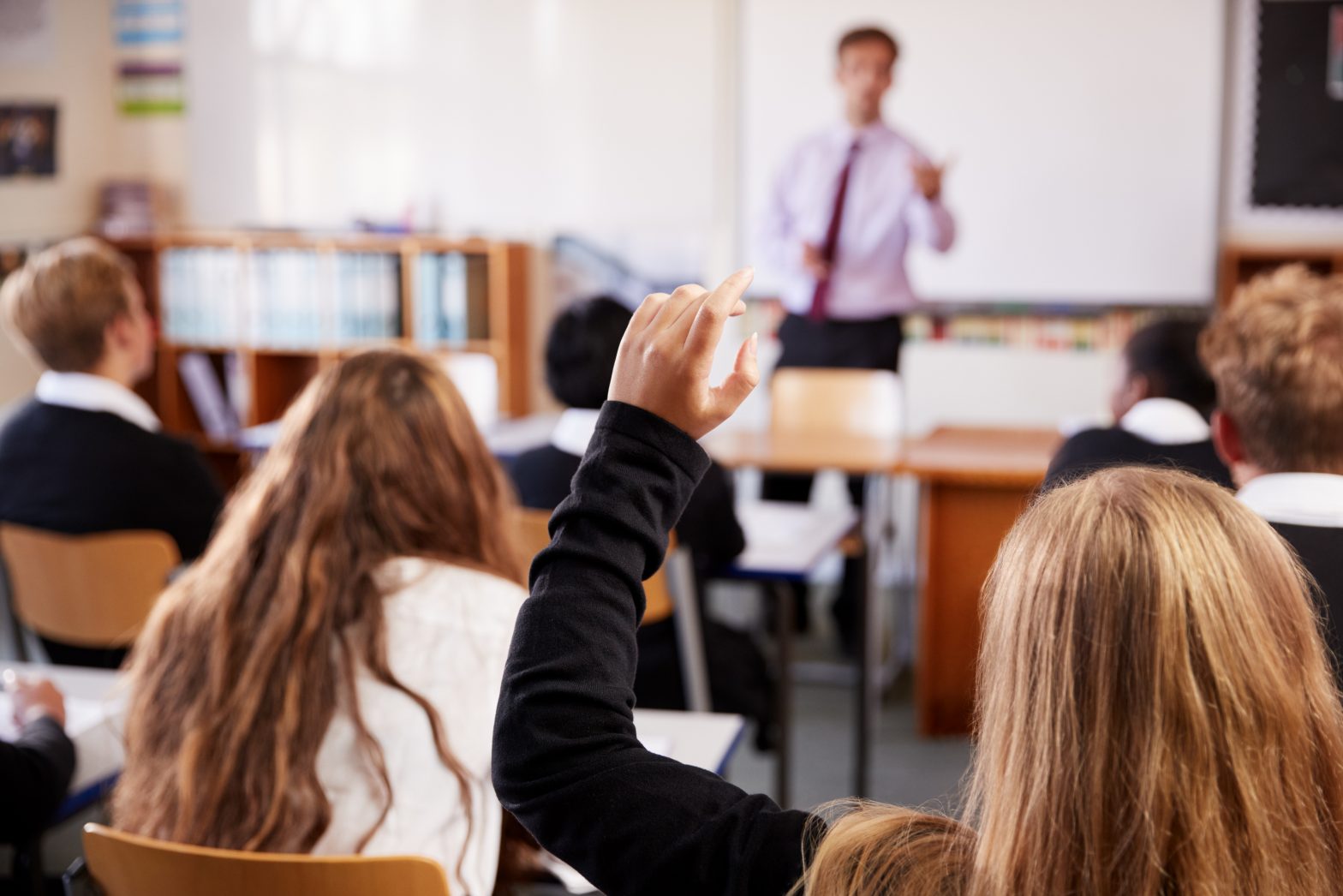 Female student in the foreground raising hand to ask question in classroom